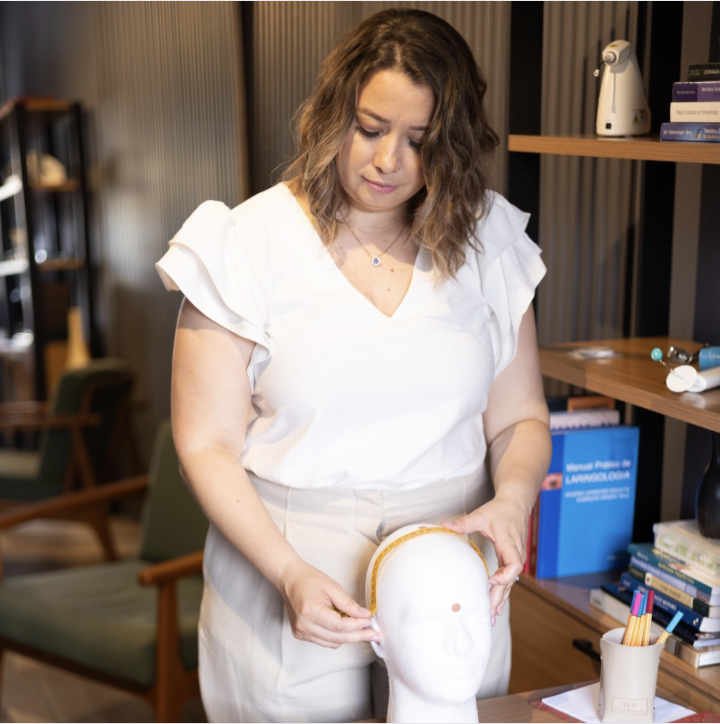 La fonoaudióloga esta en pie, detrás de una mesa, sus manos tienen una trena y está haciendo la medición de una cabeza de isopor