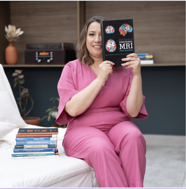 Fonoaudióloga sentada con libros a su lado y un libro en sus manos. El libro de sus manos está delante de su hemiface izquierda.