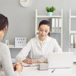 Una fonoaudióloga en su consulta, sentada en una silla. Delante hay una mesa, donde está su libreta y un ordenador. La fonoaudióloga apunta todo lo que la paciente le está diciendo. La paciente es una mujer de unos 30 años, de piel clara y pelo negro y largo