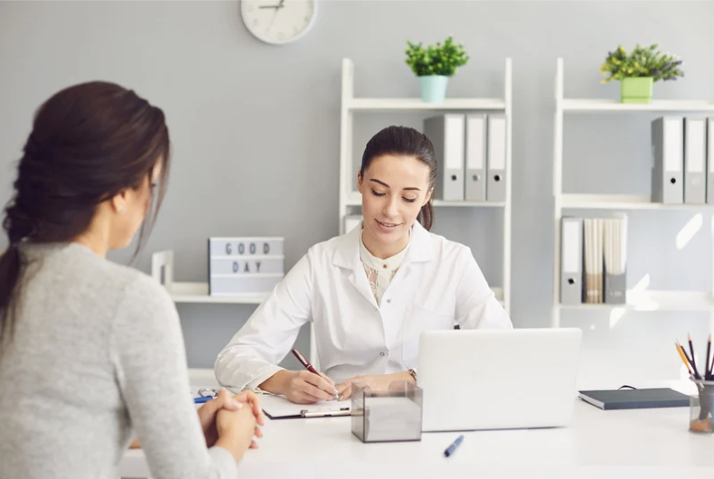 Una fonoaudióloga en su consulta, sentada en una silla. Delante hay una mesa, donde está su libreta y un ordenador. La fonoaudióloga apunta todo lo que la paciente le está diciendo. La paciente es una mujer de unos 30 años, de piel clara y pelo negro y largo
